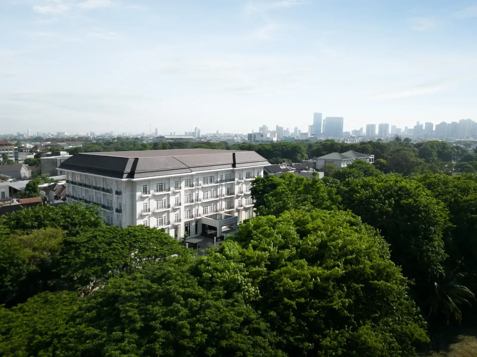 The Grand Mansion Menteng's white heritage facade above the tree canopy in Central Jakarta.