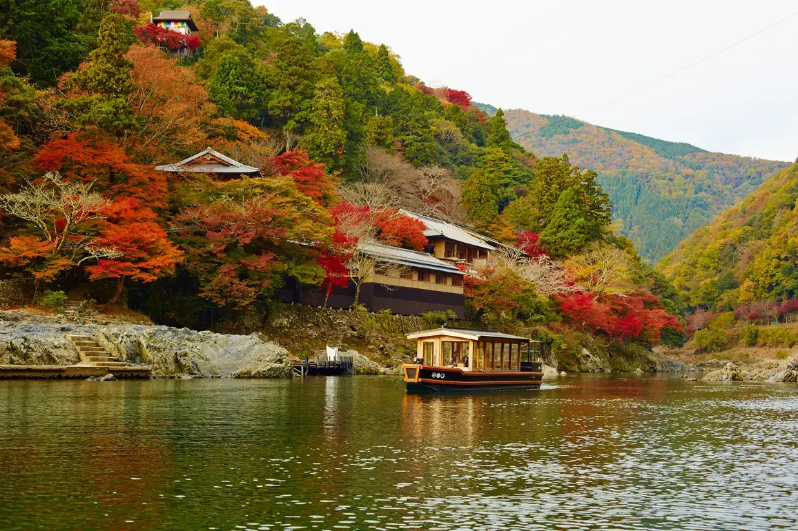 HOSHINOYA Kyoto's riverside buildings and arrival boat framed by autumn foliage in Arashiyama.