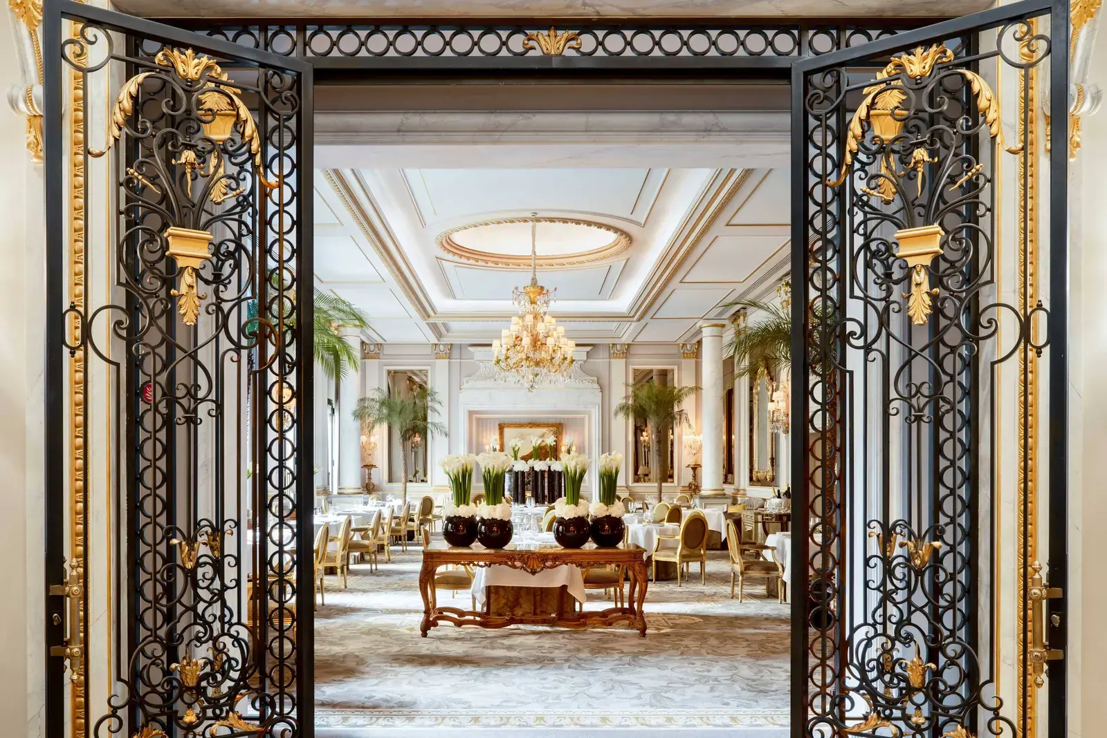 Le Cinq's dining room at Four Seasons George V, seen through gilded wrought-iron doors, with a domed gold chandelier and formally set tables beneath panelled walls
