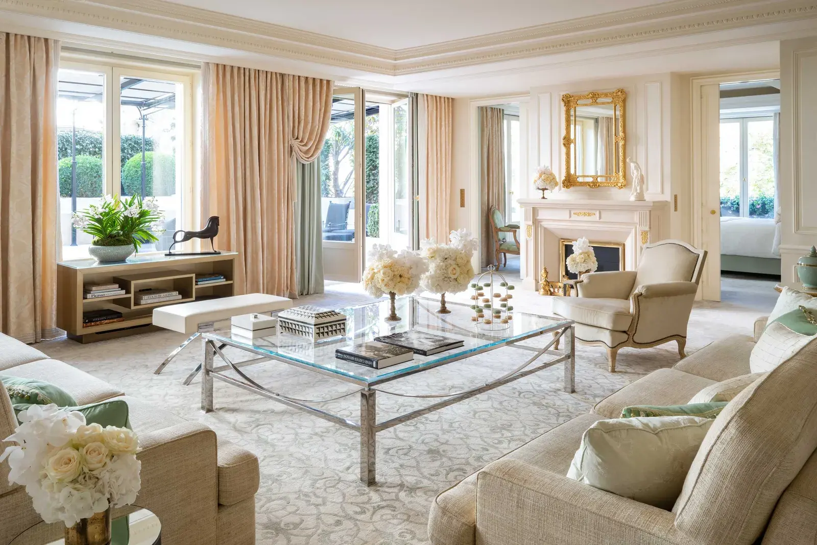 The Royal Suite living room at Four Seasons George V, with cream upholstered seating, a glass-topped table and full-height curtained windows onto a planted terrace