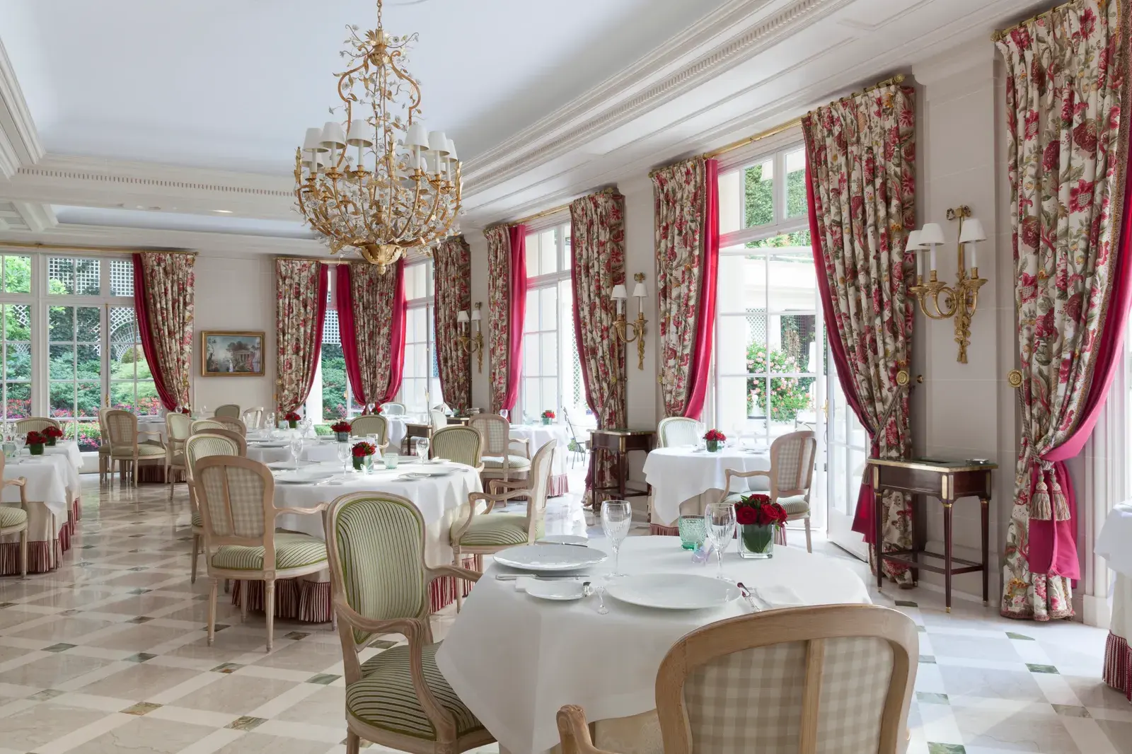 The Epicure dining room at Le Bristol Paris, with a gilt floral chandelier, crimson and floral drapery, pale marble chequerboard flooring and French windows opening onto the garden