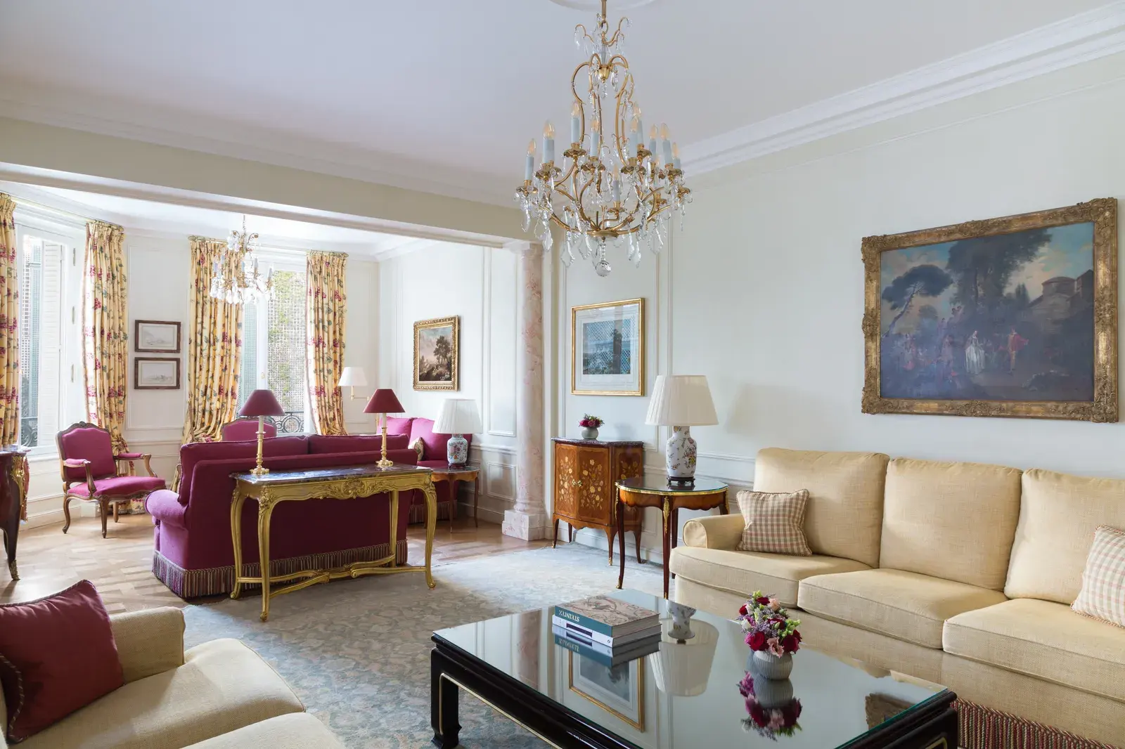 The Imperial Suite salon at Le Bristol Paris, with a crystal chandelier, raspberry and cream seating, a gilt console table and a large framed pastoral painting
