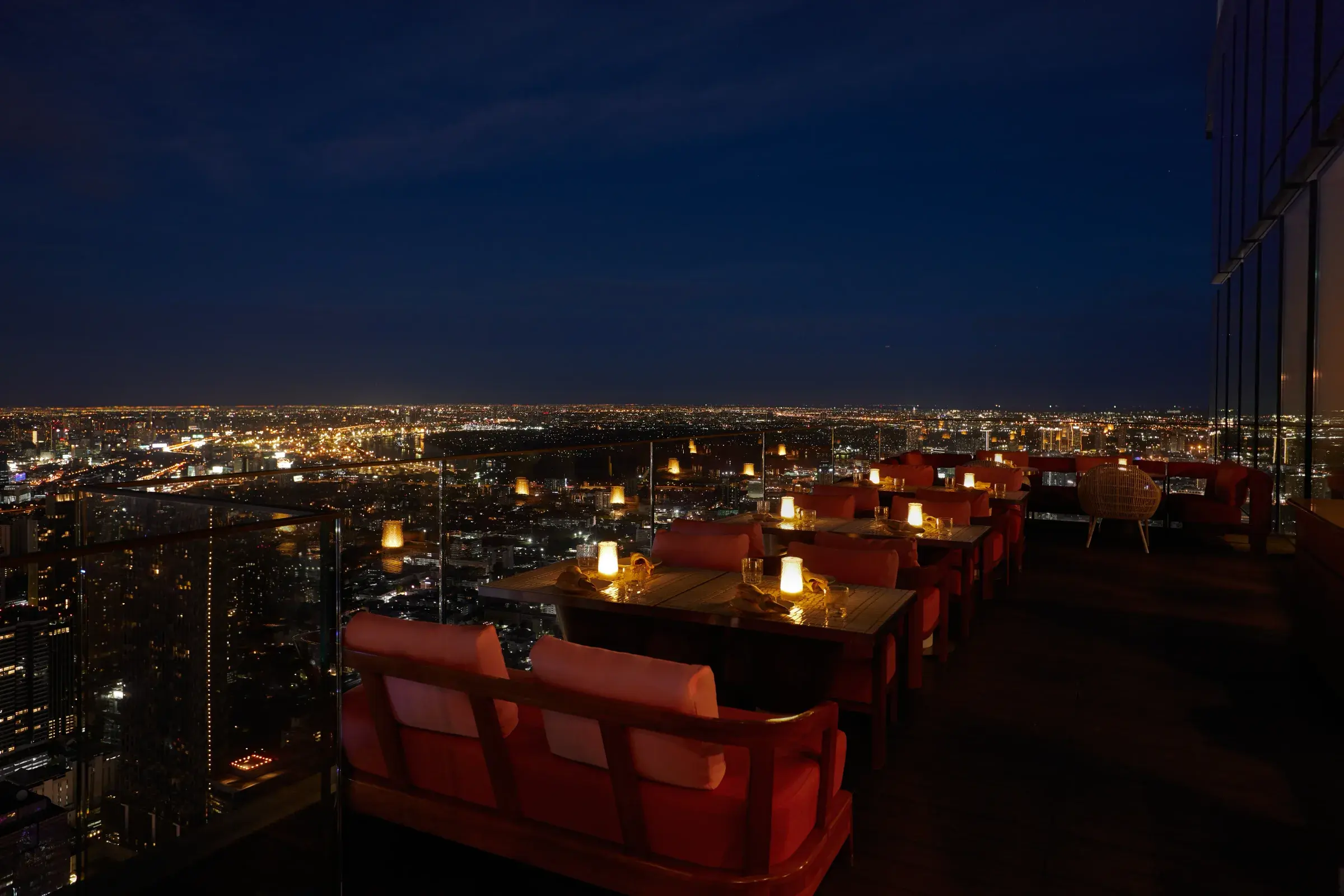 Ojo's open-air rooftop terrace at The Standard Bangkok Mahanakhon at dusk, with candlelit teak tables, coral banquettes and the lit Bangkok skyline below